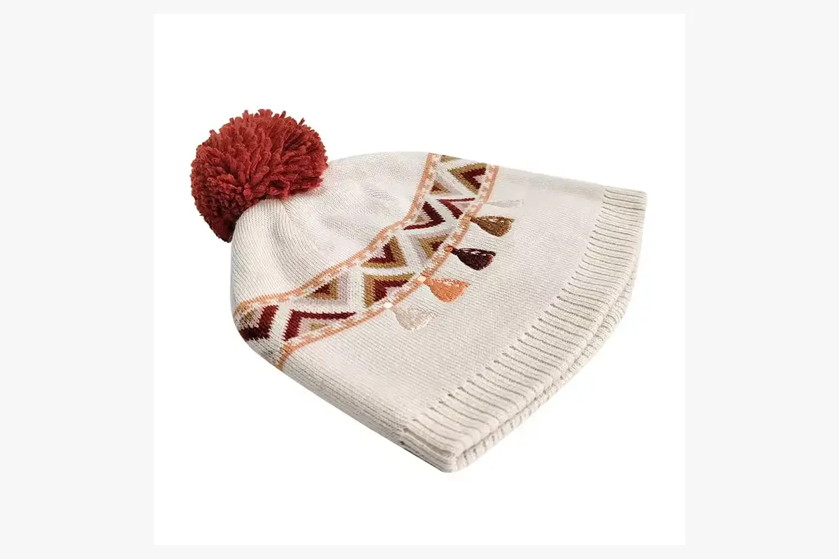Cream pom pom beanie shown at an angle with a red geometric knitted band