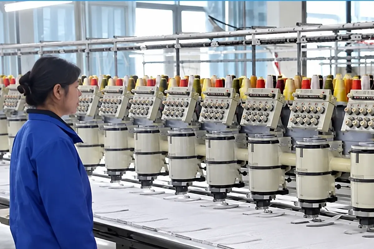 Factory operator overseeing a row of industrial knitting machines and yarn cones