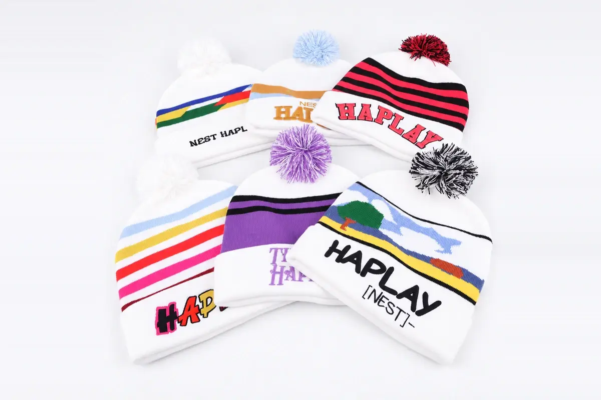 Six white pom-pom beanies with colorful stripes, patterns and HAPLAY branding