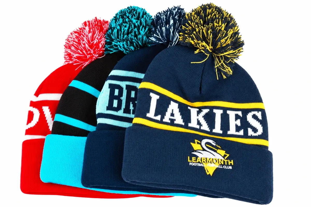 Four colorful pom-pom beanies with striped cuffs and team-style lettering.