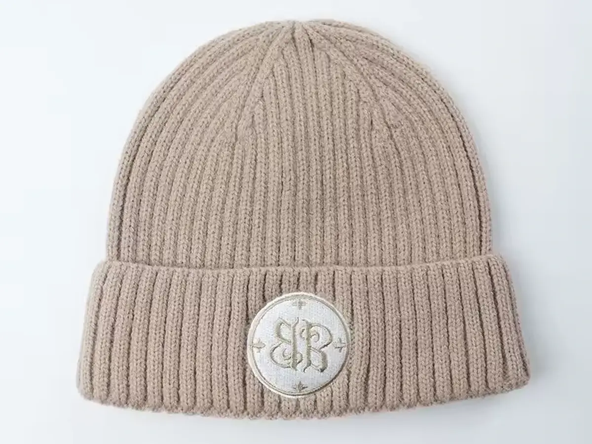 Beige rib-knit beanie with a folded cuff and small front badge.