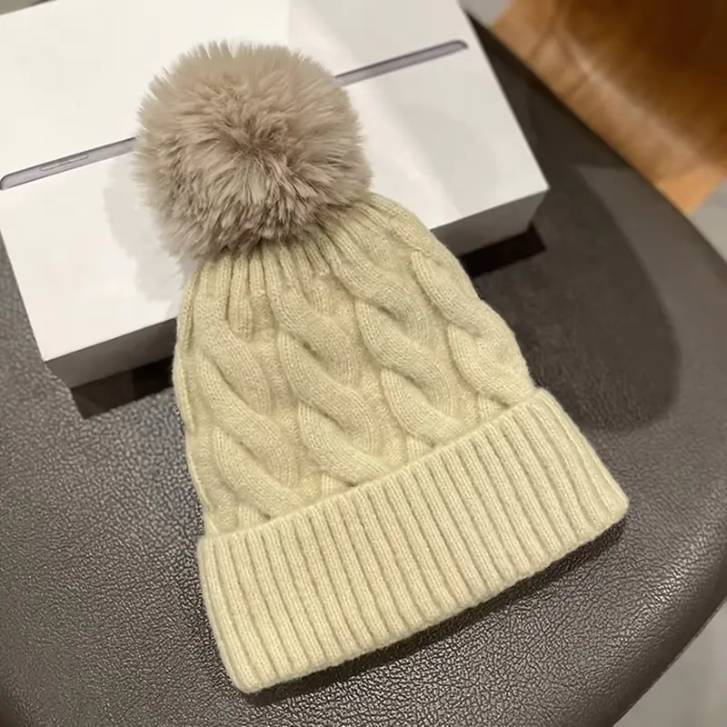 Beige cable-knit beanie showing raised cables, folded cuff and pom pom