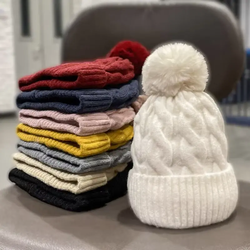 Several cable-knit beanies in different colors beside a cream pom beanie